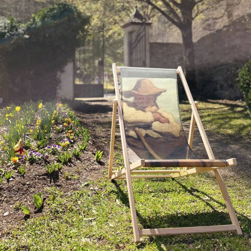 Ein Liegestuhl im Garten mit einem Kunstmotiv und bunten Blumen im Hintergrund. Frühlingsoase im Freien.