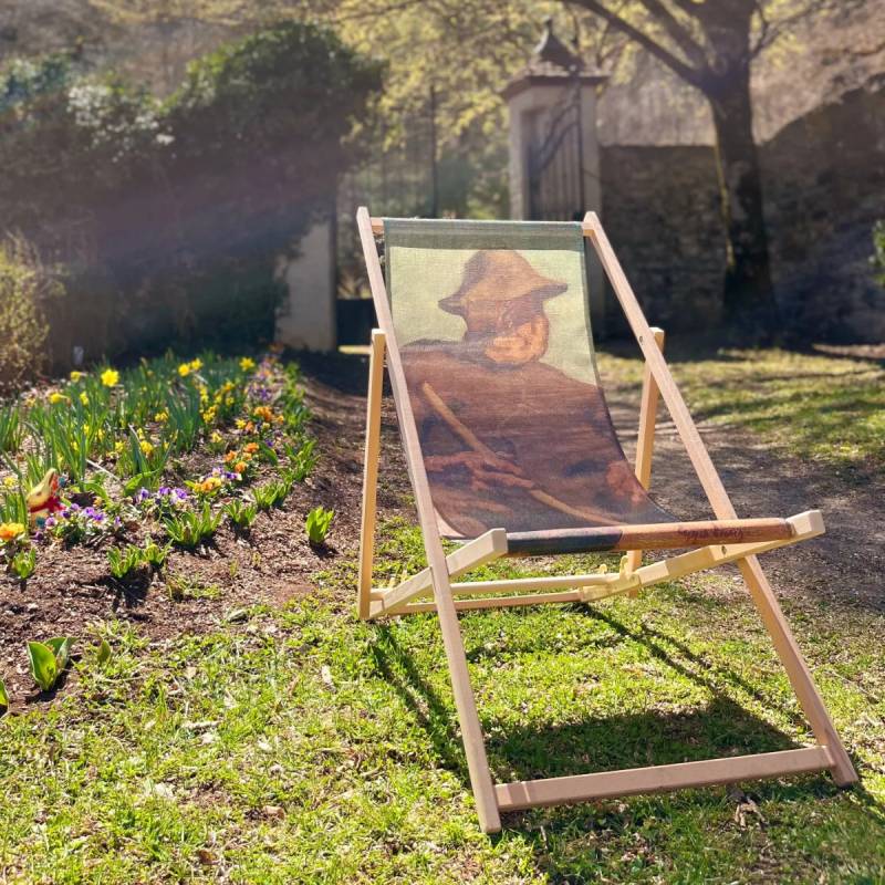 Ein Liegestuhl im Garten, bedruckt mit einem Bild eines Mannes, umgeben von bunten Blumen und sonnigem Licht.